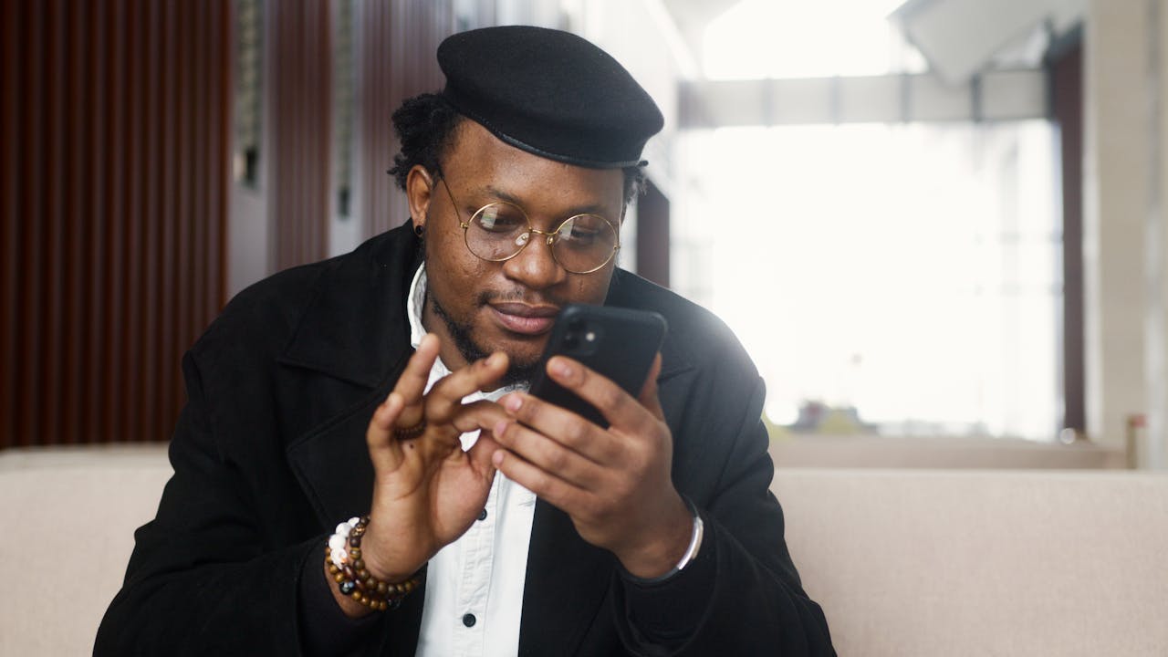 A cheerful man wearing a stylish outfit uses a smartphone while seated in a modern indoor setting.